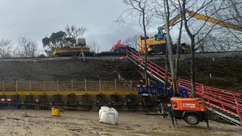 A view from the ground of where the landslip was and the embankment has been rebuilt, with machinery and people in hi-viz suits on train tracks.