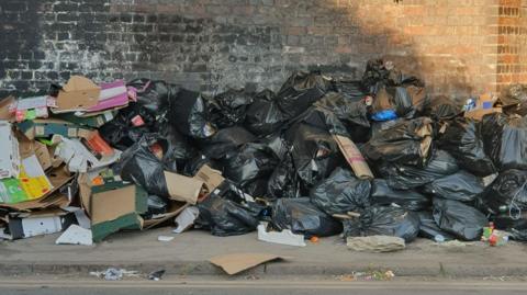 Piles of black bin bags, cardboard and other litter against a dark wall near a kerbside. There are about 30 bin bags in the picture. 