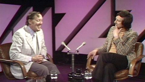 Bill Haley sits in a chair, opposite Barry Norman, in front of a purple background