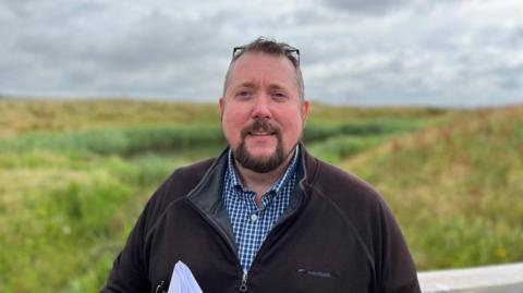 A man in a black jumper and check shirt smiling. He is in a field.