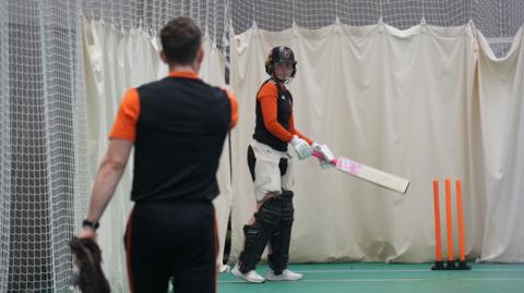 Grace Ballinger stands with bat in hand listening to a coach give her feedback in training. She has full batting kit on and is in indoor nets at Trent Bridge.