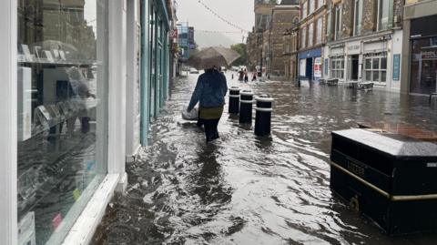 A person walks down Winchester High Street carrying and umbrella, with flooding water up to her knees