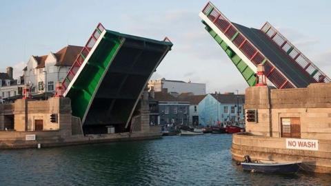 Green and red bridge in the up position in two halves - buildings stand along a harbourside to the left of the bridge.