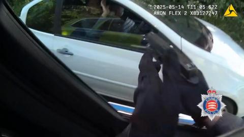 A close-up of someone's hands wearing black gloves, holding a black gun, pointing at a white car.