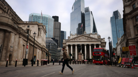 A lone man in light brown puffer jacket walks across the street in front of the old stock exchange in Bank. The Bank of England's stone building is visible on the top left while a 26 red double-decker bus drives in the direction of the man to the right