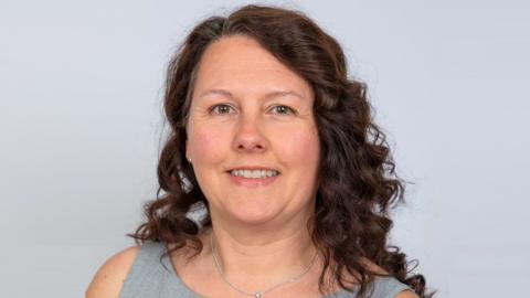 A woman with long, wavy brown hair is wearing a grey sleeveless top. She is looking at the camera and has a grey background behind her. 