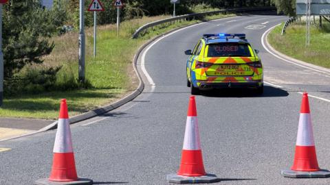 Orange cones are blocking off the dual carriageway, with a sign in the rear window of a police car reading: "Closed".