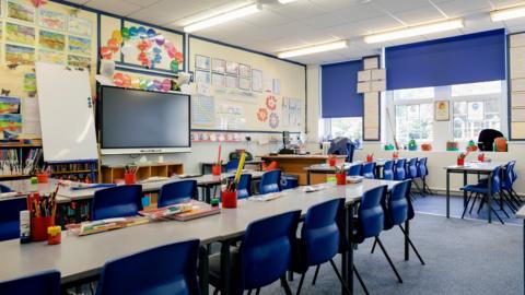 A bright, empty classroom with desks, chairs and pencils in the foreground