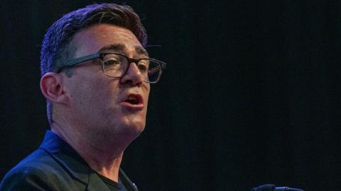 Andy Burnham wearing a black jacket with a black t-shirt talking to an audience with a black background behind him