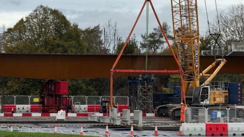 A tall yellow crane is operating next to a large steel bridge. The area on the ground underneath the bridge is blocked off with temporary construction fencing. 