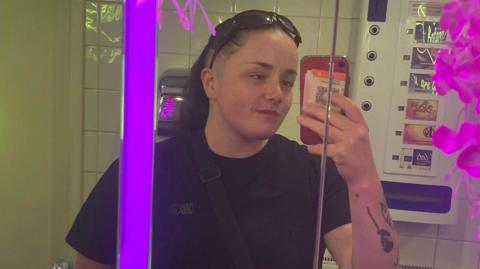 Amber Stebbings wearing a dark T-shirt, taking a photo of herself in the mirror with a red phone. She has tattoos on the arm she is holding the phone up with. In the background is a wall-mounted vending machine. 