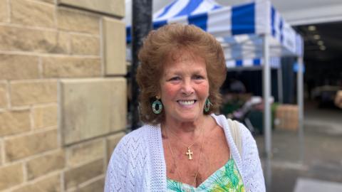 A woman with a white cardigan smiles. Behind her, a market is blurred.