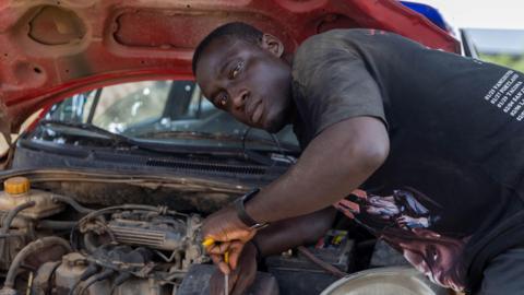 Nathaniel Qainoo, dressed in black, fixes the engine of a car