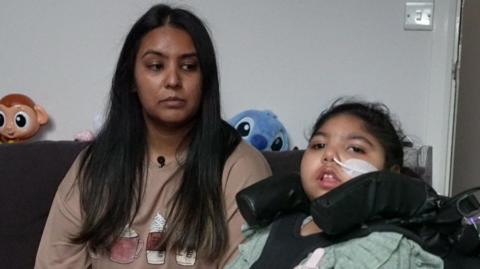 A picture of mum Namla (left) with dark brown hair and daughter Sara (right) sitting in a wheelchair with a feeding tube in her nose.
