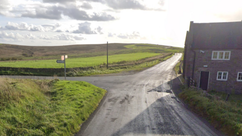 A country road leading to a junction, with a cottage to the left. Acres of hills and green farmland can be seen ahead
