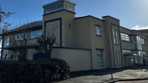 The outside the of the Isle of Man Airport terminal building, which is cream coloured with blue trim and large glass windows.