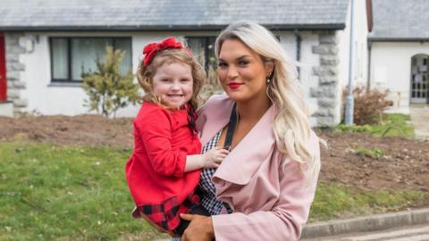 Natalie McLaughlin holds her young daughter Ella in her arms. She is smiling, has long blonde hair, and is wearing silver earrings, a pink overcoat and a black and white checked blouse. Ella is also smiling, and has a red bow in her long brown hair. The little girl is wearing a red dress. Behind them is a garden and house.