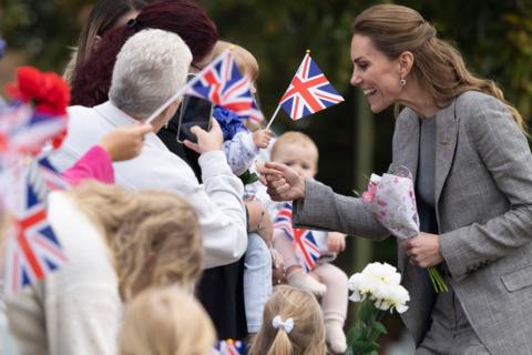 The Princess of Wales, wearing a grey suit and holding a small bunch of flowers, smiles as she meets a croud of people who are waving flags and taking photographs.