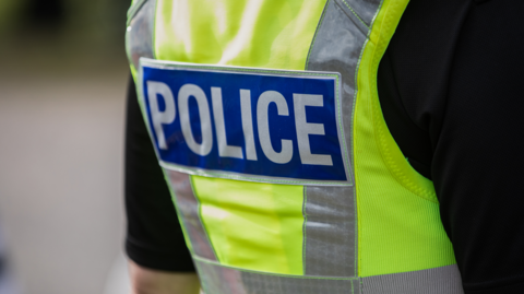 A close-up of the back of a police officer, wearing a green high-viz police jacket