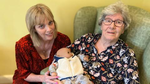 Two women looking at the camera and smiling. The woman on the right is sat in a green armchair and is cradling a baby doll. The woman on the right is crouched down next to her.
