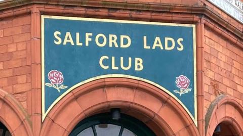 A green sign with gold letters reading 'Salford Lads Club' placed over the red brick arches that adorn the entrance to the club. Green wooden doors can be seen beneath the arches, with a street sign for 'Coronation Street' placed on the left hand side of the building. 