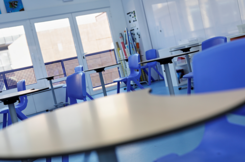 A generic classroom with a number of blue chairs sat at single-person desks. On the wall there is a blank white board. 