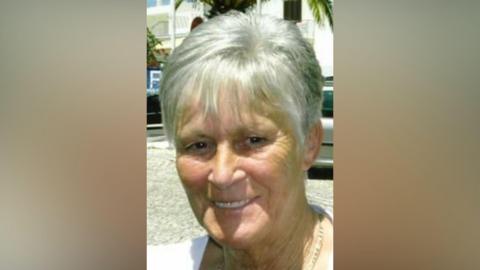 June Sanderson has a short grey bob haircut and a necklace on. She is smiling.