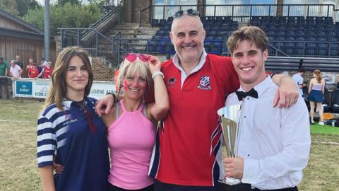 John, his wife Tina, daughter Georgina, and son Sean. They are stood on a rugby pitch, hugging for a family photo. John wears a red and black rugby strip, and has sunglasses on top of his head. Georgina wears a blue and white sports shirt and Tina is wearing a pink sleeveless top. Sean is holding a trophy and is wearing a white shirt with a black bow tie.