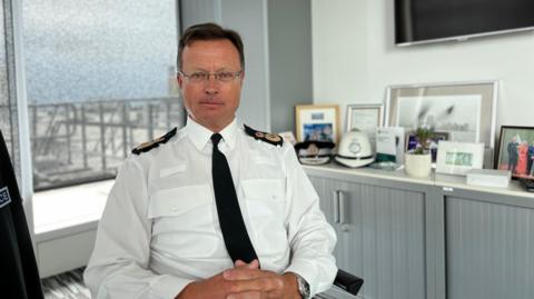 Officer Smith looks at the camera wearing his uniform and sitting in a chair while behind him are photos and and police hats on a filing cabinet