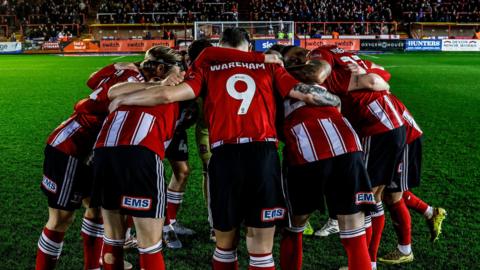 Exeter City team huddled together in front of a stand full of fans. The kits are red and they are all wearing black short.
