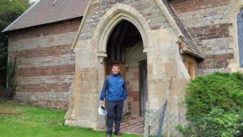 Sam Pratley stands in the doorway of the disused Church of St Andrew, Wolferlow. He is wearing a blue jacket and has a white helmet in his hand. The arched doorway and building is made of stone and there are tiles on the floor in the entrance.