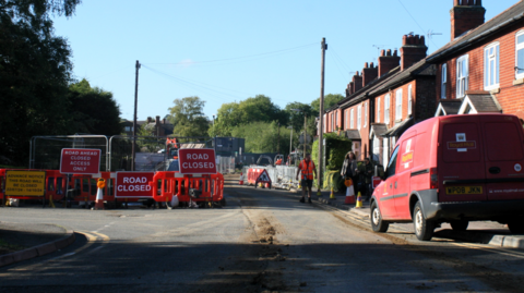 Roadworks across Brook Road. Barriers and fences are across the road with red signs saying Road Closed. There is a row of terraced homes to the right of the road and a red Royal Mail van parked near them.  