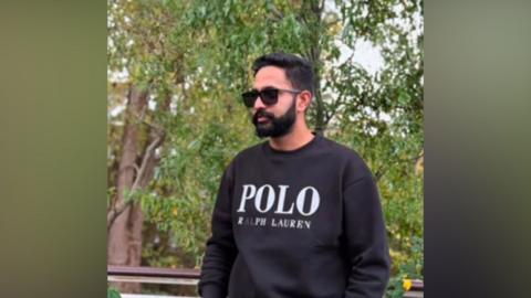 A young man standing outdoors near some trees. He is wearing a dark jumper and sunglasses and has a black beard and black hair.