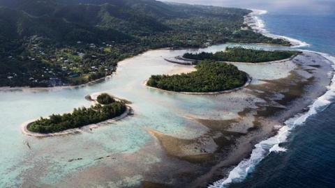 Aerial shot of the Cook Islands. Waves are crashing onto the shore, where there are three small islands filled with bushy green trees.