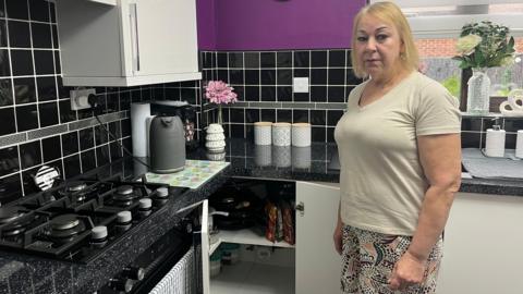 A woman with short blonde hair looking straight-faced and standing in her purple-tiled kitchen in Swindon with white units and black worktops, with the smart meter on view in an open cupboard.