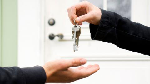 A person holding silver keys above another person's hand. They are in front of a white door with a light green frame.