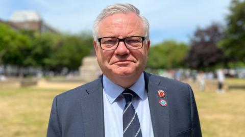 Constable Richard Vibert stood in a green space wearing a blue suit and tie and looking straight at the camera. He is wearing glasses. There are trees and people blurred behind him.