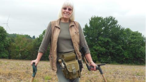 Nettie Edmondson is standing outside, in front of trees, with grass behind her, holding a spade and a metal detector in each hand. She has shoulder-length blonde hair, with a fringe, is wearing glasses, and has on a gilet, a beige top and trousers with a tool belt around her middle. 