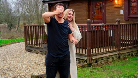 Stacey Solomon covers Joe Swash's eyes in front of a wooden cabin.