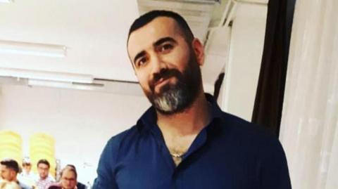 Erdal is smiling at the camera, wearing a blue shirt with the top couple of buttons open. He has short dark brown hair and a dark brown beard that is slightly grey around his chin. 