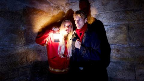 Daisy May and Charlie Cooper stand in the corner of a dark, old, building, Daisy is holding up a lantern and they are both wearing frightened and concerned expressions on their faces. 