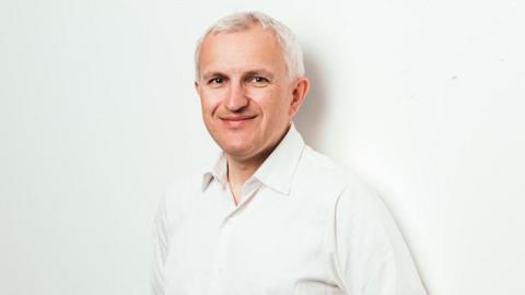 A white man with white hair in a white open neck shirt. He is smiling at the camera 