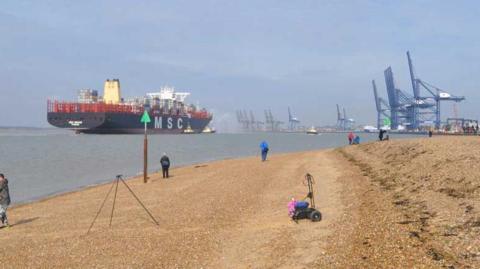 A container ship approaches the Port of Felixstowe on the left of the picture. The cranes dominate the horizon on the right. A pebbled beach is in the foreground with a few people on it.