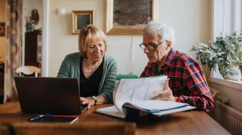 A woman with blonde short hair is sitting in front of a laptop. She is sitting next to a man wearing a red and blue plaid shirt and glasses. He is reading from a booklet.