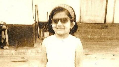 Pragna Hay as a young girl in Uganda with headband on, wearing sunglasses, and wearing white dress. The photo is old, a little blurry and sepia-toned.