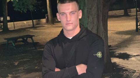 Ben Moncrieff wearing a black hoodie with a yellow logo on the upper left sleeve. He has short brown hair and is posing for a picture with his arms folded. It is dark and in the background you can see an open park area with wooden picnic benches and trees. 