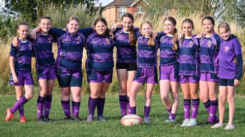 Ten girls, all under the age of 12, stand in a line on a grass pitch in their striped purple jerseys with their arms around each other's shoulders. They smile at the camera, with a rugby ball on the grass in the middle of them.