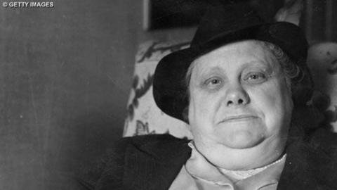 Older woman wearing black bowl hat sits on patterned chair for photograph.