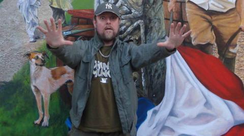 A man with a beard, wearing a New York Yankees cap, a jacket, brown shirt and chain around his neck, has his arms outstretched in front of him in front of a painting.