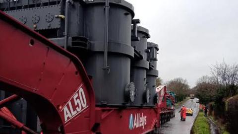 Grey machinery moving along a road on a red trailer. Two men in orange overalls are standing near the trailer. A police car is driving in front of the load. The trailer is marked with letters and numbers - AL50 - and the word "Allelys".